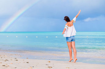 A young woman joyfully walks along a beach with a rainbow on the horizon.