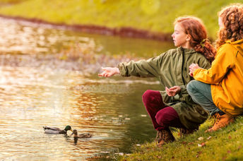 Two young girls feed ducks at a pond.