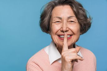 A smiling woman holds her finger to her lips, signaling for quiet.