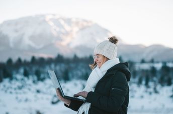 A young smiling woman uses a laptop outdoors in the mountains.