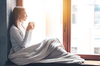 A woman sits by a window and holds a cup.