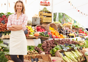 A vendor stands in front of her vegetable stand at an outdoor market.