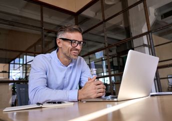 A man wearing glasses smiles while looking at a laptop screen.