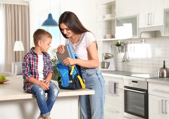A smiling mother helps her son pack a backpack.