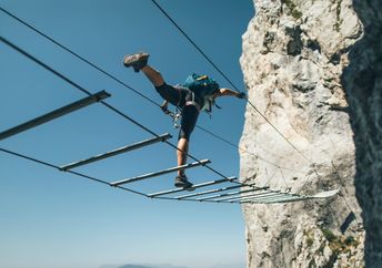 A person balances on a metal rope bridge against a blue sky and mountain.