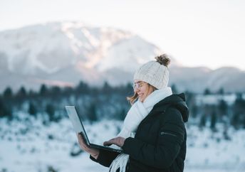 A young smiling woman uses a laptop outdoors in the mountains.