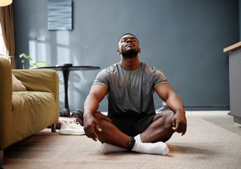 A young adult man sits cross-legged on a carpet in a living room.