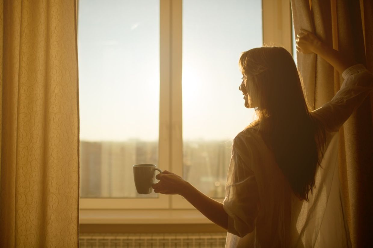 Woman in the morning. Attractive sexy woman with neat body is holding a cup with hot tea or coffee and looking at the sunrise standing near the window in her home and having a perfect cozy morning.
