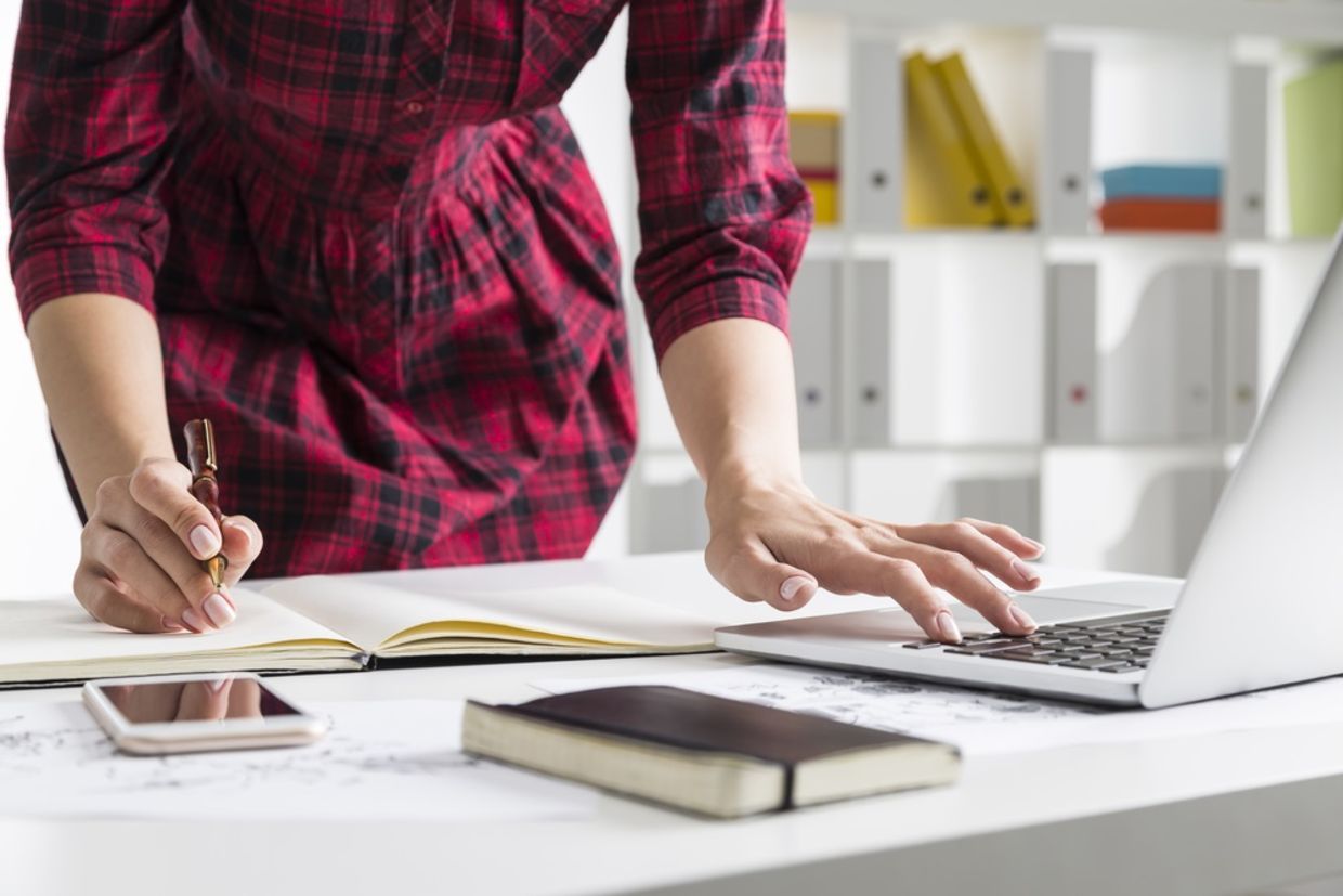 Close up of woman in checkered dress typing on her laptop while standing and making notes