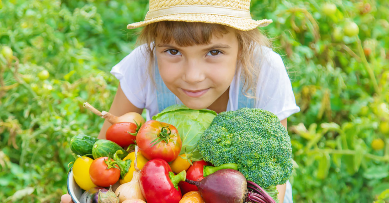 A young girl in a field holds a basket filled with freshly-picked vegetables.