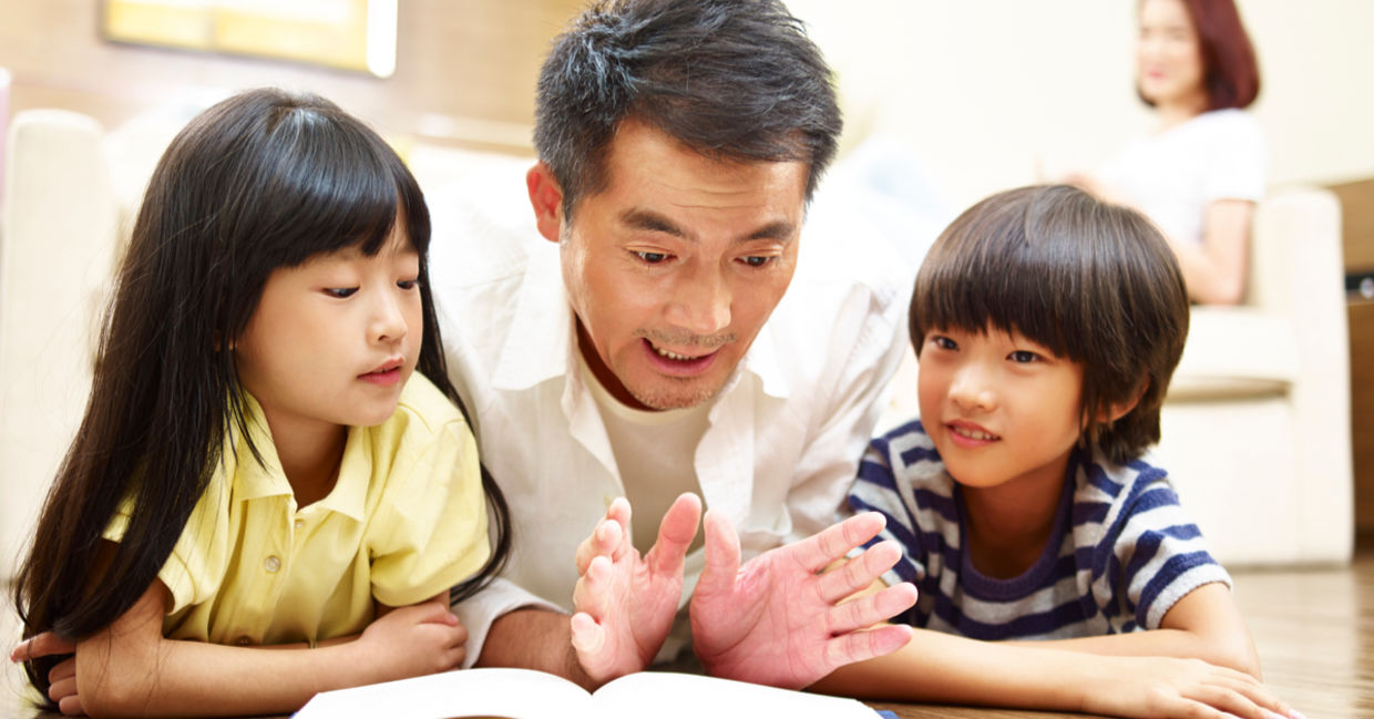 A father lies on the floor reading a book to his two children.