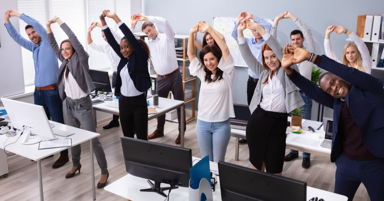 A group of office workers stand and stretch at their desks.