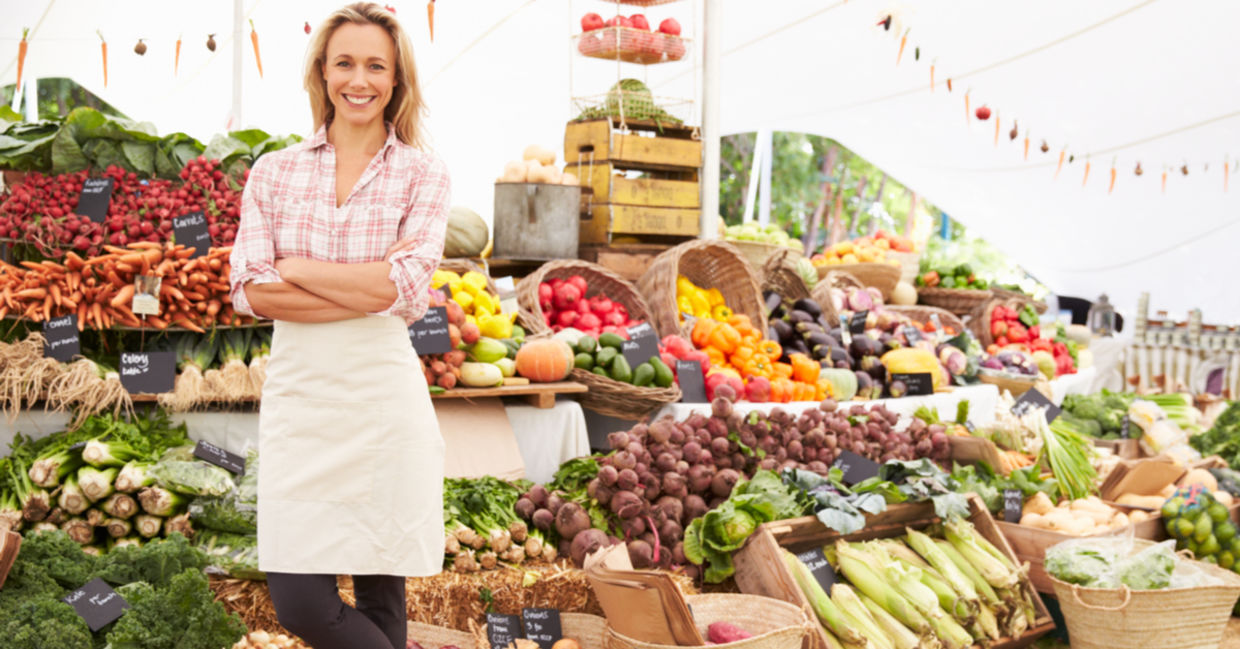 A vendor stands in front of her vegetable stand at an outdoor market.