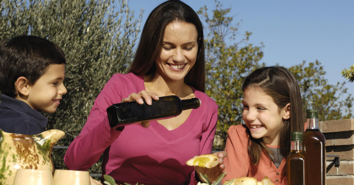 A mother with two young children pours olive oil onto bread.