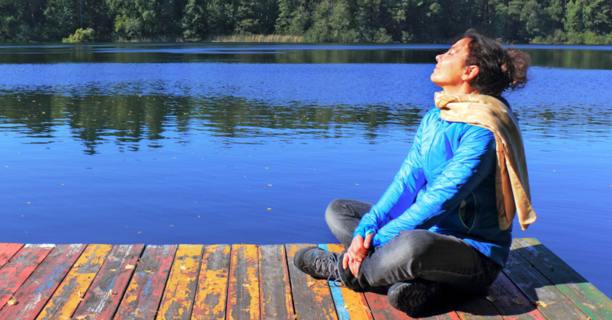A woman sitting on a dock by a lake is doing a breathing exercise to help tone her vagus nerve.