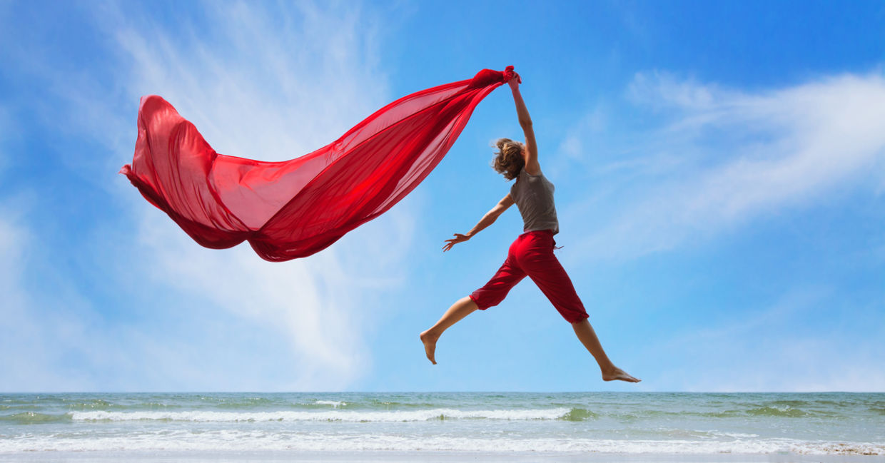 A happy, fulfilled woman jumps on the beach, trailing a bright red scarf high in the air.