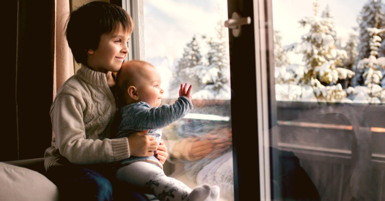 Boy holding his baby brother, sitting by the window in living room, looking at a snowy landscape outdoors.