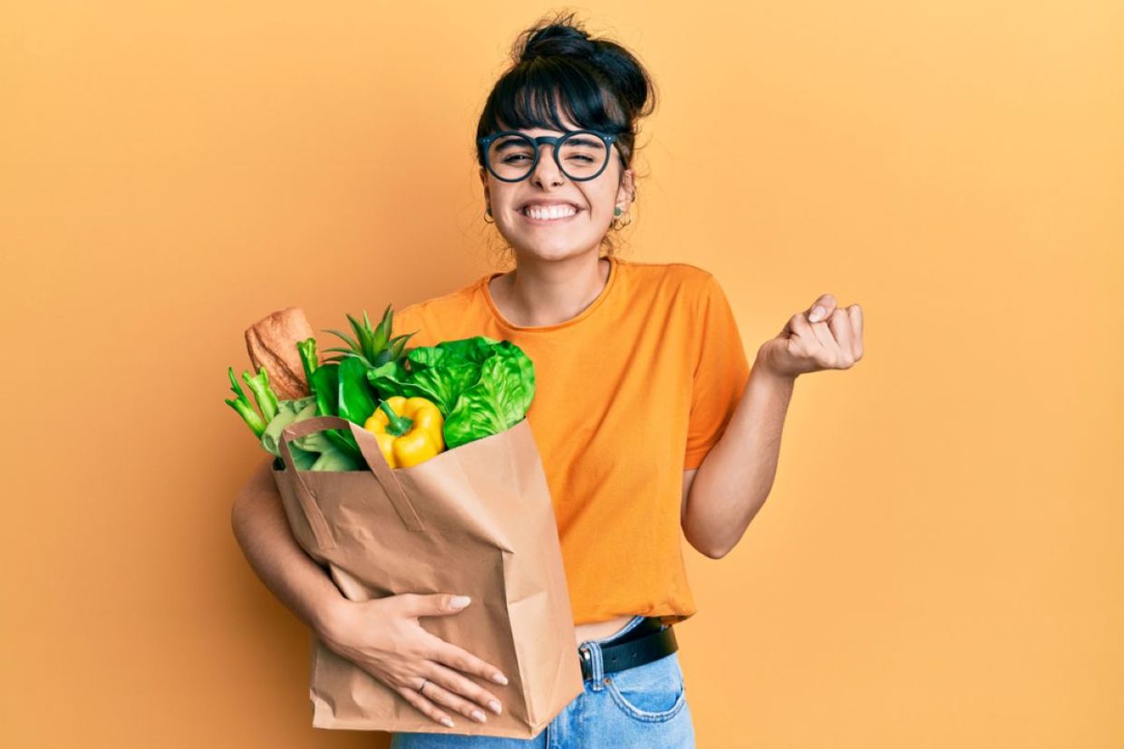 A woman smiles and holds a bag filled with food.