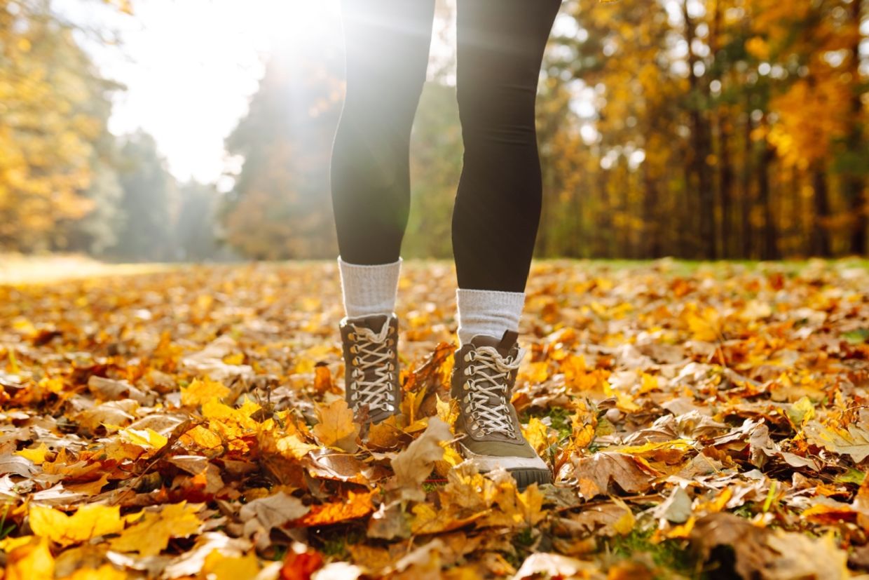 A woman wears hiking boots and stands on autumn leaves in a park.