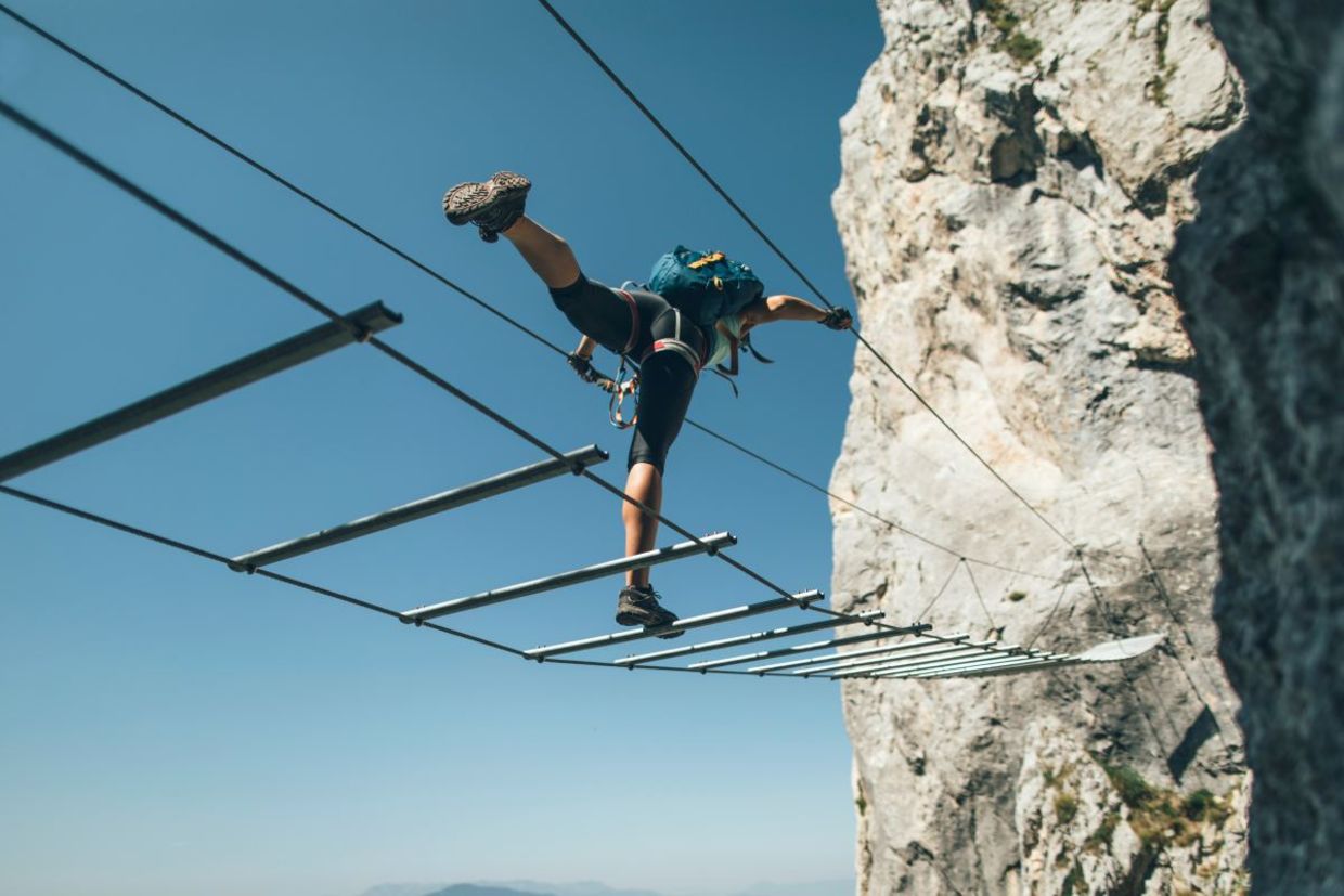 A person balances on a metal rope bridge against a blue sky and mountain.