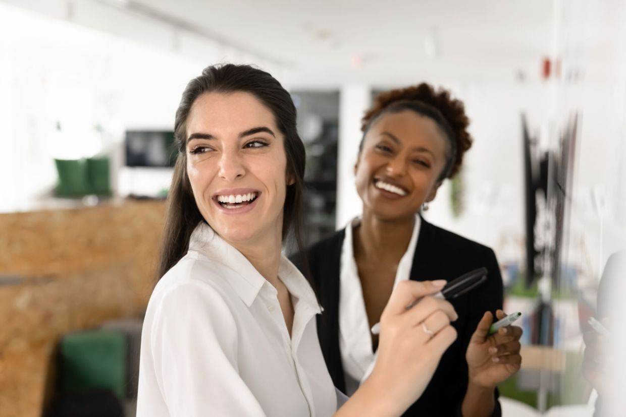 Two female coworkers smiling as they prepare to write on a whiteboard.