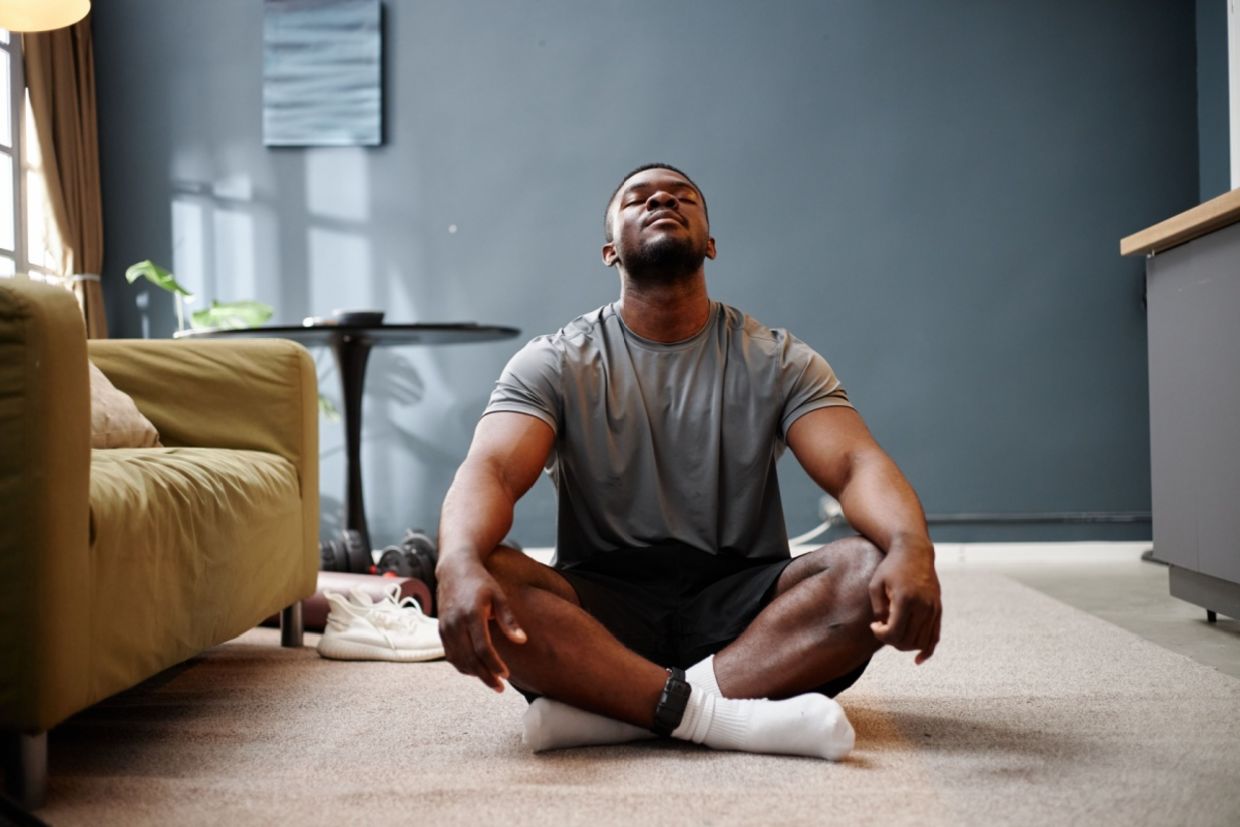 A young adult man sits cross-legged on a carpet in a living room.