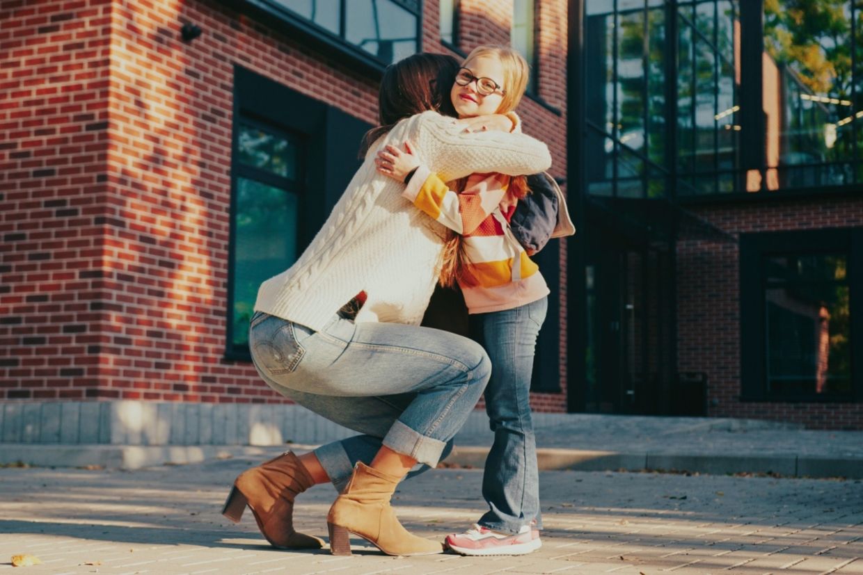 A mother kneels down to hug her daughter.
